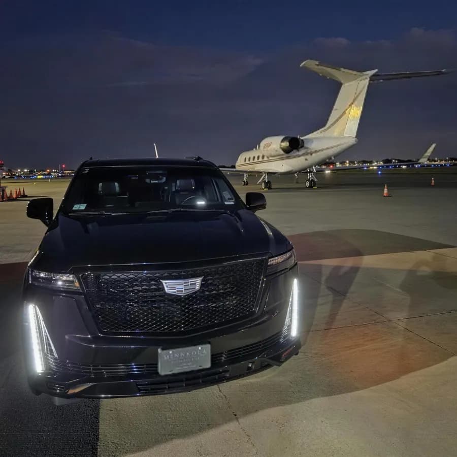 Black Cadillac Escalade luxury SUV stationed next to a private jet on a terminal tarmac at night.